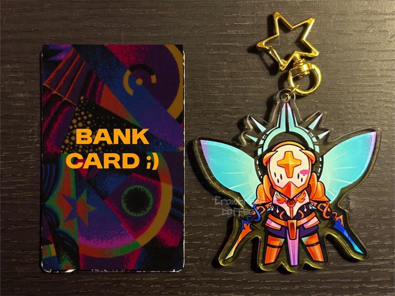 May include: A colorful bank card with the text "BANK CARD :)" next to a charm featuring a character with wings, a gold star, and a gold clasp. The charm has orange, blue, and purple accents.