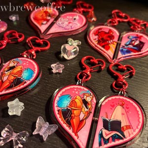 Puede incluir: Llaveros rosas en forma de corazón con cierres rojos en forma de corazón. Cada llavero está dividido por la mitad, con un personaje de dibujos animados diferente en cada lado. Hay dijes y accesorios transparentes adicionales esparcidos alrededor de los llaveros.
