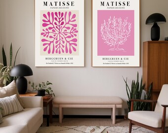 May include: Two framed art prints featuring the name "MATISSE" and "PAPIERS DÉCOUPÉS". One print has a pink floral design on a beige background, the other has a white tree design on a pink background. The prints are displayed in a living room setting.
