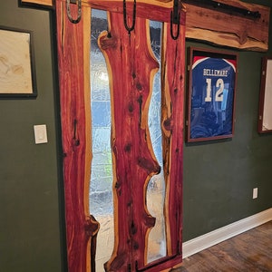 May include: A wooden sliding door with a large glass panel featuring a unique, organic design. The door is made of reddish-brown wood with a natural grain pattern. The glass panel is clear and has a wavy, irregular shape. The door is mounted on a black metal track.