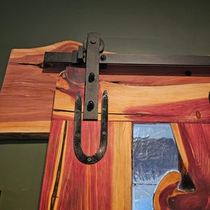 May include: A wooden door with a rustic, natural finish. The door has a large, metal, U-shaped handle and a black metal track for sliding. The door has a clear glass panel with a wood grain pattern.