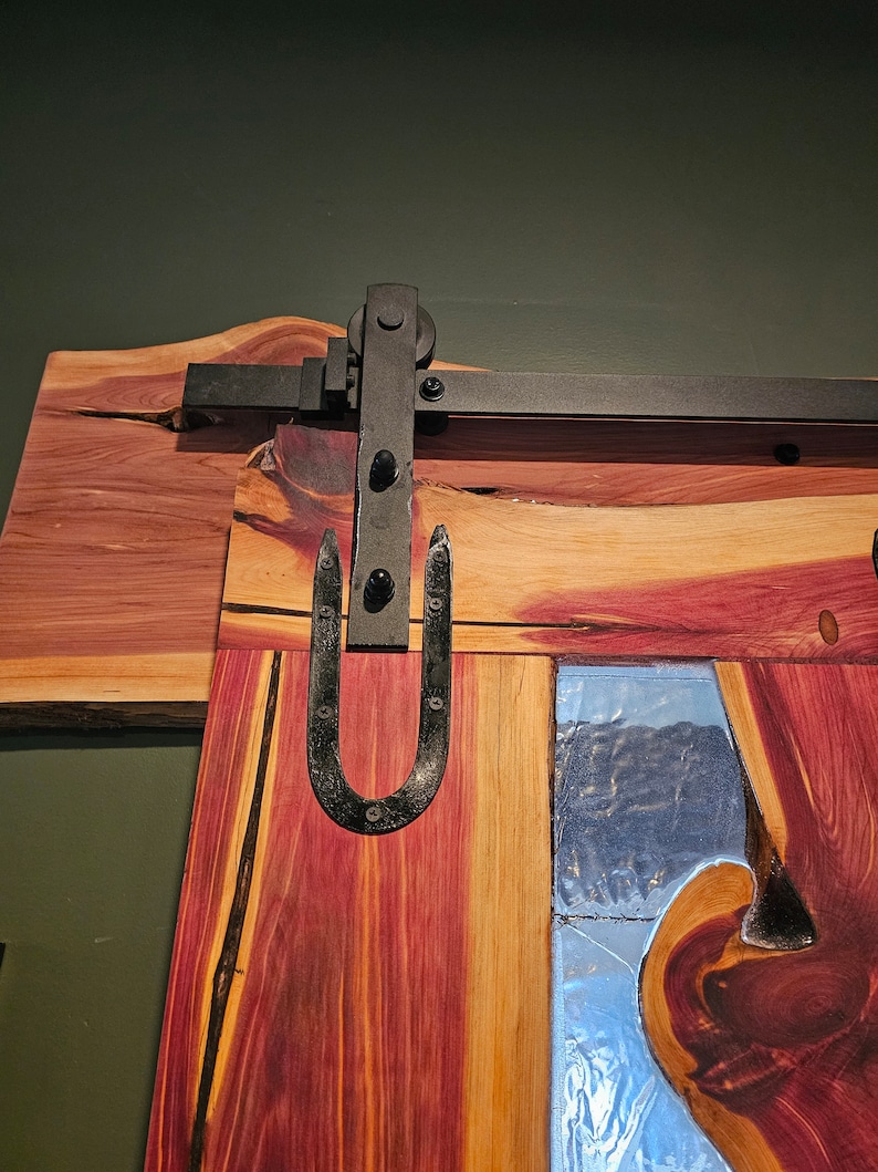 May include: A wooden door with a rustic, natural finish. The door has a large, metal, U-shaped handle and a black metal track for sliding. The door has a clear glass panel with a wood grain pattern.