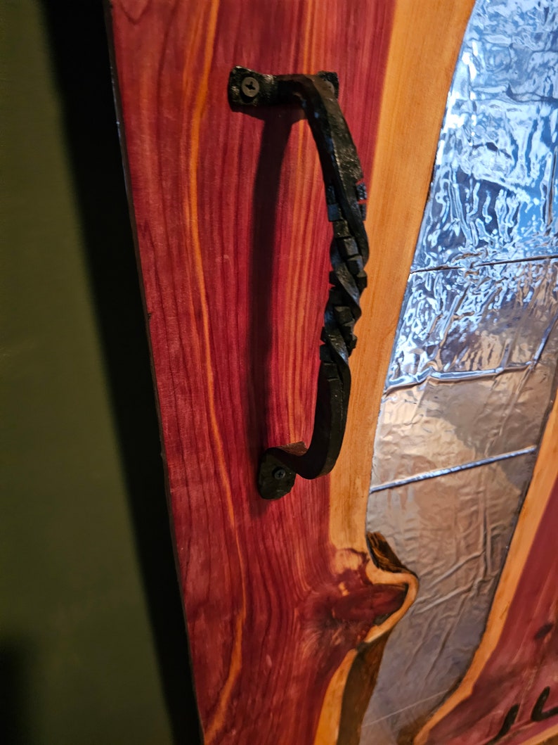 May include: A close-up of a wooden door with a black metal handle. The door is made of a dark wood with a reddish hue. The handle is twisted and has a decorative design. The door is partially open, revealing a window behind it.