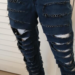 May include: Dark blue denim jeans with multiple horizontal rips and frayed edges. The jeans are decorated with small black rhinestones.