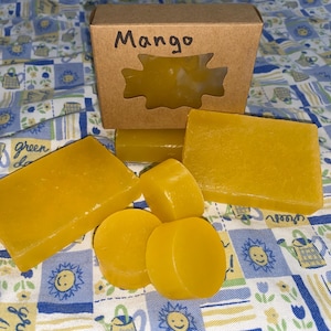 以下が含まれることがあります： 黄色い石鹸がいくつかあり、長方形と円形です。模様のある布の上に配置されています。「Mango」と書かれた小さな段ボール箱もあり、石鹸が見えるように切り取られています。