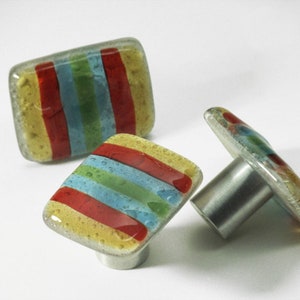 Op de afbeelding: Drie kleurrijke glazen kastknoppen met een zilveren metalen basis. De knoppen hebben een gestreept patroon in rood, geel, blauw en groen.