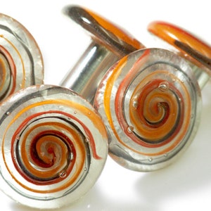 Può includere: Quattro tappi per bottiglia in vetro con un motivo a spirale arancione, marrone e argento. I tappi sono rotondi e hanno una base in metallo argentato.