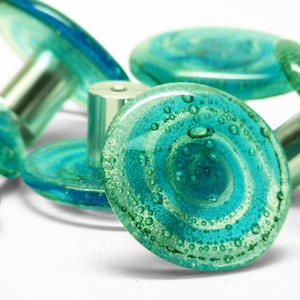 May include: A collection of decorative glass cabinet knobs. Each knob features a swirling teal and turquoise design with a silver metal stem. The glass has a bubbly texture, and the knobs are arranged on a white surface.