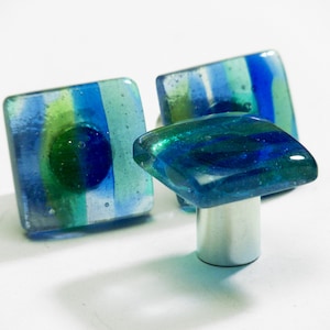 May include: Three square glass cabinet knobs with an abstract design in blue, green and white. The knobs have a silver metal base.