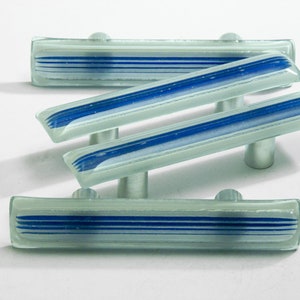 Peut inclure: Quatre poignées de meubles en verre de forme rectangulaire. Chaque poignée a un corps en verre transparent avec des rayures bleues et blanches et une poignée en métal argenté. Les poignées sont disposées sur un fond blanc.