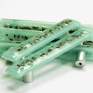 Puede incluir: Conjunto de cuatro tiradores de armario de vidrio azul aguamarina con una placa de montaje de metal plateado. El vidrio tiene un aspecto texturizado y moteado.