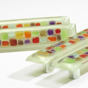 Può includere: Due pomelli per armadi in vetro con un design a mosaico che presenta quadrati colorati in tonalità di rosso, arancione, giallo, verde, blu e viola. I pomelli sono traslucidi e hanno una tinta verde chiaro.