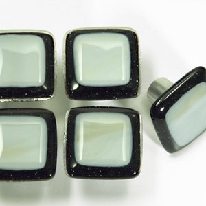 Puede incluir: Cinco pomos de armario cuadrados de vidrio con un centro blanco y un borde negro. Los pomos tienen un acabado negro moteado.