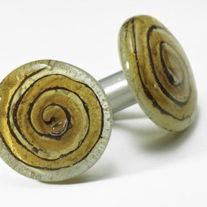 May include: A pair of cufflinks with a clear glass design. Each cufflink features a golden-yellow spiral pattern with dark brown lines. The cufflinks are connected by a silver-colored metal bar.