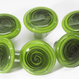 Puede incluir: Seis pomos de armario de vidrio verde con un diseño negro en espiral. Los pomos son redondos y tienen una base de metal plateado.
