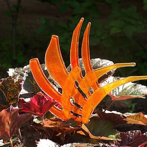 Könnte beinhalten: Orangefarbene Glasskulptur in Form einer stilisierten Blume mit mehreren Blütenblättern. Die Skulptur steht in einem Beet aus roten und grünen Blättern.