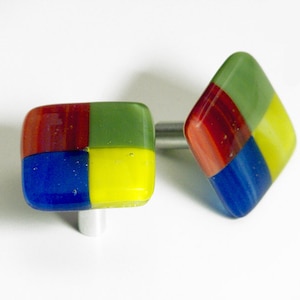 Op de afbeelding: Twee vierkante glazen kastknoppen met een geometrisch ontwerp in rood, groen, blauw en geel. De knoppen zijn bevestigd aan zilveren metalen bases.