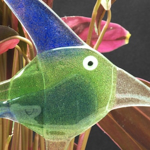 May include: A glass hummingbird feeder with a green and blue iridescent finish. The feeder has a clear beak and a white eye.