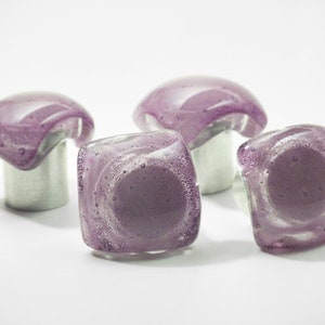 Puede incluir: Cuatro pomos de armario de vidrio morado con bases plateadas. Los pomos tienen una superficie texturizada y burbujeante.