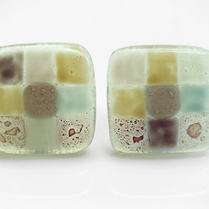 Könnte beinhalten: Ein Paar quadratische Glasohrringe mit Mosaikdesign. Die Ohrringe sind aus klarem Glas mit braunen, gelben und blauen Akzenten.