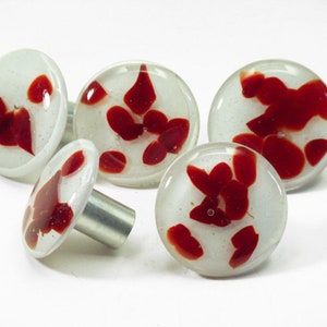 Könnte beinhalten: Eine Sammlung von fünf dekorativen Schrankknöpfen. Jeder Knopf hat ein klares, abgerundetes Glasdesign mit roten, abstrakten Formen, die in einen weißen Hintergrund eingebettet sind. Die Knöpfe haben einen silberfarbenen Metallstiel zur Montage.