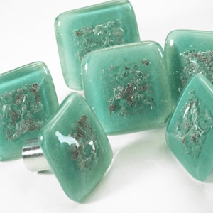Puede incluir: Un juego de seis pomos de armario cuadrados y en forma de diamante. Los pomos son de color turquesa translúcido con un diseño texturizado de color plateado en el centro. Cada pomo está unido a una base metálica plateada.