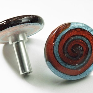 Puede incluir: Dos pomos redondos de cristal para armario con un poste de metal plateado. Un pomo tiene un patrón de espiral negro, azul y rojo. El otro pomo tiene un patrón de espiral negro, rosa y blanco.