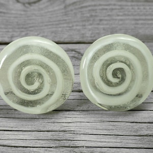 Op de afbeelding: Een paar ronde, witte glazen oorbellen met een spiraalvormig ontwerp. De oorbellen zijn doorschijnend en hebben een lichtgroene tint.