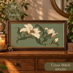 Könnte beinhalten: Ein rechteckiges Kreuzstichmuster mit weißen Lilienblüten und dunkelgrünen Blättern auf salbeigrünem Hintergrund, gerahmt in einer dunklen Holzleiste. Das Bild enthält den Text PDF Pattern und Cross Stitch 100x150.
