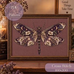 Könnte beinhalten: Ein gerahmtes Kreuzstichmuster mit einer Libelle, deren Flügel in Violett, Creme und Gold mit Blumenmotiven, Mondsicheln und Sternen auf dunkelmalvenfarbenem Grund verziert sind. Der Text PDF Pattern und Cross Stitch 100x200 ist sichtbar.