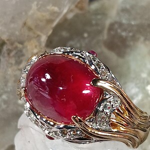 May include: A gold and silver ring featuring a large, oval-shaped, deep red gemstone. The gemstone is surrounded by a halo of smaller, clear stones. The ring's band has an intricate, flowing design.