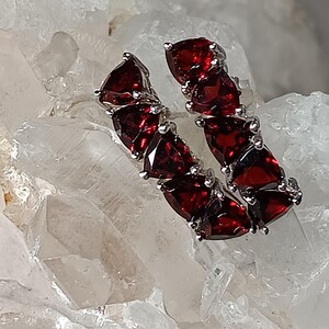 May include: A pair of silver earrings featuring a series of vibrant, triangular, deep red gemstones. The earrings are arranged in a linear design, showcasing the rich color and sparkle of the stones. The earrings are displayed on a textured, light-colored surface.