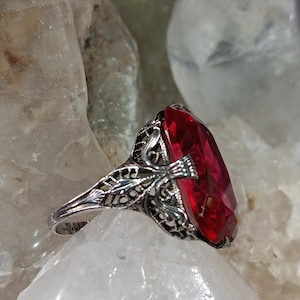 May include: A sterling silver ring with a large, faceted, rectangular red gemstone. The ring features intricate filigree detailing around the gemstone and along the band. The ring is set on a white, crystalline surface.