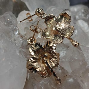 May include: A pair of gold-colored flower-shaped earrings. The earrings have a post-back closure and are detailed with textured petals and a small, round stud at the center. The earrings are displayed on a light-colored, crystalline surface.