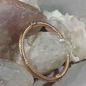 May include: A gold-colored, twisted rope design bracelet. The bracelet is a bangle style, with a clasp closure. The jewelry is displayed on a bed of clear and purple crystals.