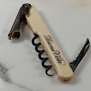 May include: Ivory-colored wine opener with a stainless steel corkscrew, bottle opener, and knife. The words "Buena Vista" are printed in cursive on the handle. The tool is designed for opening wine bottles.