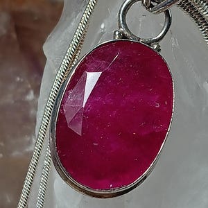 May include: An oval-shaped, deep magenta-red gemstone pendant set in a polished silver-toned metal bezel. The pendant hangs from a silver-toned snake chain necklace, featuring a faceted surface that reflects light across its vibrant, saturated color.