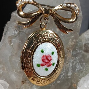 May include: A gold-toned vintage brooch featuring a bow with faux pearls and a locket pendant. The locket has a white enamel oval with a red rose and green leaves. The brooch is a classic accessory.