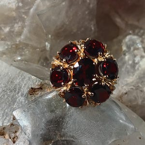 May include: A gold-toned ring featuring a cluster of deep red gemstones. The gemstones are round and set closely together, creating a floral or cluster design. The ring is set against a light, textured background.