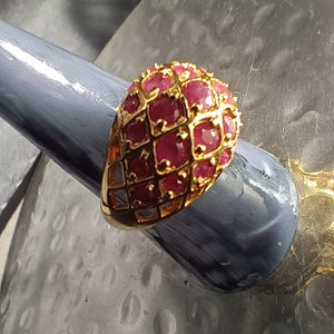 May include: A gold ring with a large, domed setting featuring a cluster of red ruby stones. The ring has a delicate, openwork design.