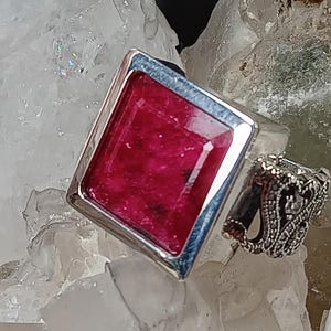 May include: A silver ring with a large, rectangular, deep red gemstone. The ring is set in a silver band with a contrasting blue border. Another smaller, ornate silver ring is visible in the background.
