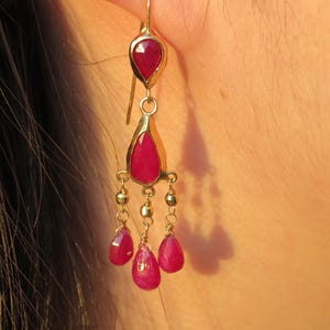 Peut inclure: Une boucle d'oreille chandelier en or avec un rubis en forme de goutte en haut, relié à une structure centrale portant trois gouttes de rubis pendantes et de petites perles dorées. La boucle est suspendue à un crochet en or et est rouge vif.