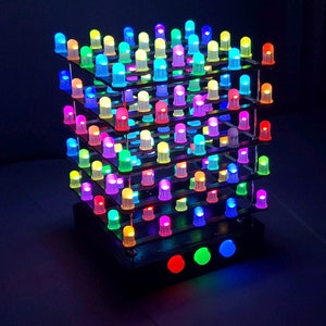 Puede incluir: Una pantalla de luz LED en forma de cubo con múltiples niveles, cada uno con numerosas luces LED multicolores. Las luces están dispuestas en un patrón de cuadrícula, emitiendo colores vibrantes. La base presenta tres botones iluminados.