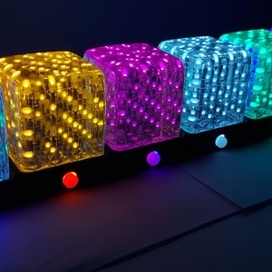 May include: A series of six illuminated cube-shaped LED lights in a spectrum of colours: blue, yellow, purple, green, blue, and red. Each cube sits on a black base with a matching coloured button. The lights are arranged in a row, creating a vibrant display.