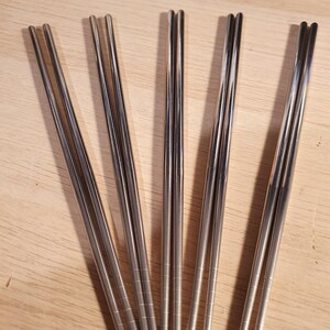 May include: A set of six silver chopsticks, arranged in a fan shape on a wooden surface.