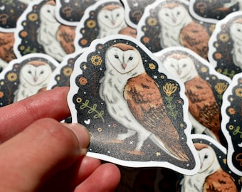 May include: A sticker featuring a white and brown barn owl with a black background. The owl is surrounded by small yellow and orange flowers and stars. The sticker is printed on white paper.