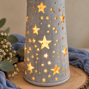 Könnte beinhalten: Eine zylindrische graue Lampe mit glatter, matter Oberfläche und zahlreichen sternförmigen Ausschnitten in verschiedenen Größen. Die Lampe strahlt ein warmes, goldenes Licht aus und beleuchtet die Sternmuster auf der grauen Außenfläche.