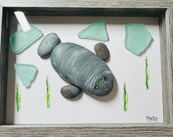 May include: A framed art piece featuring a hand-painted manatee made of stone. The manatee is gray with painted details, surrounded by sea glass and painted seaweed. The frame is gray.