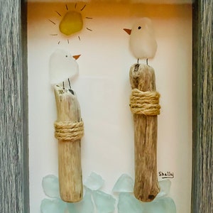 May include: A framed artwork featuring two white glass birds perched on driftwood posts, with a yellow glass sun and blue glass sea glass at the bottom. The artwork is signed "Shelly".