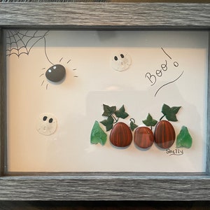 May include: Framed Halloween artwork with stone-crafted elements: a spider, ghosts, and pumpkins. The pumpkins are brown and red, with green leaves. The word "Boo!" is written in black. The frame is grey.